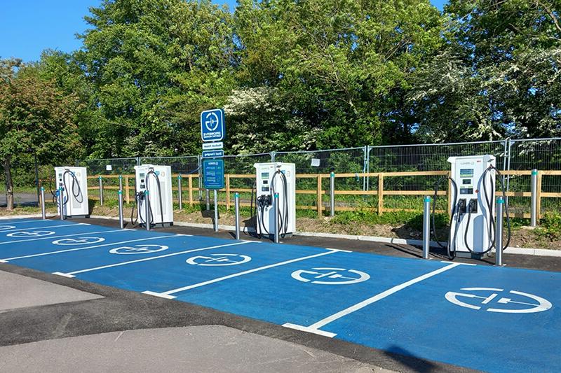 A line of four charging stations for electric vehicles, white boxes with black cables, with blue parking spaces in front of them. A wooden fence and green trees can be seen in the background