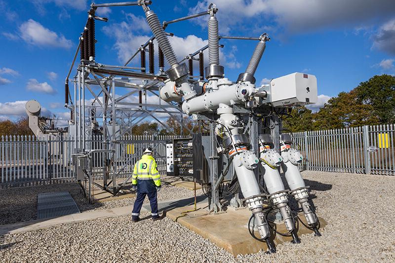 An electrical substation with wires and coils in a metal structure, with a gravel floor and a metal surrounding fence. A G2 Energy worker in high-vis and hard hat is in the middle